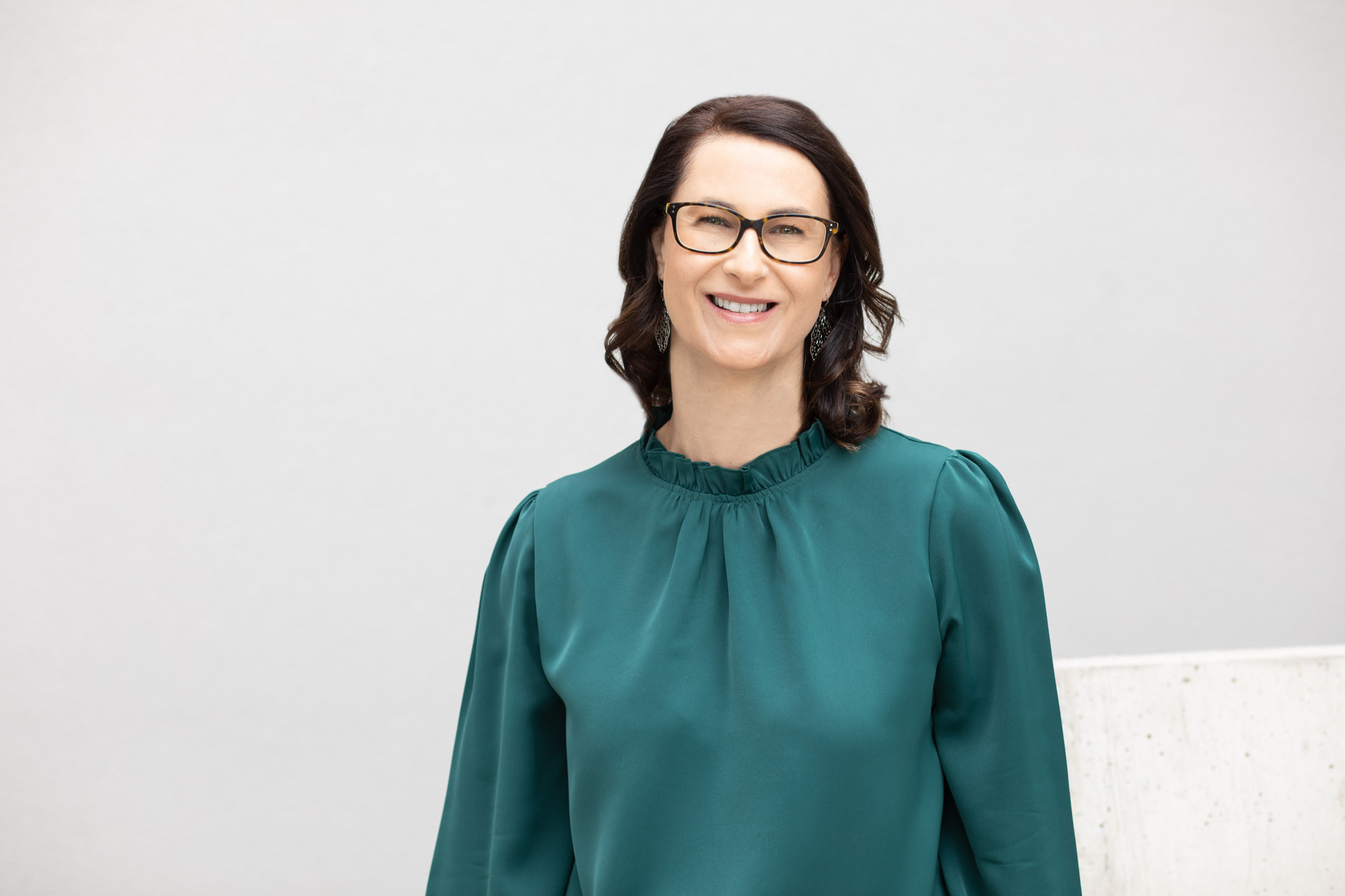Eine Person mit schulterlangem, gelocktem Haar trägt eine grüne Bluse vor einem neutralen, hellgrauen Hintergrund. Das Gesicht ist verpixelt und unkenntlich gemacht.