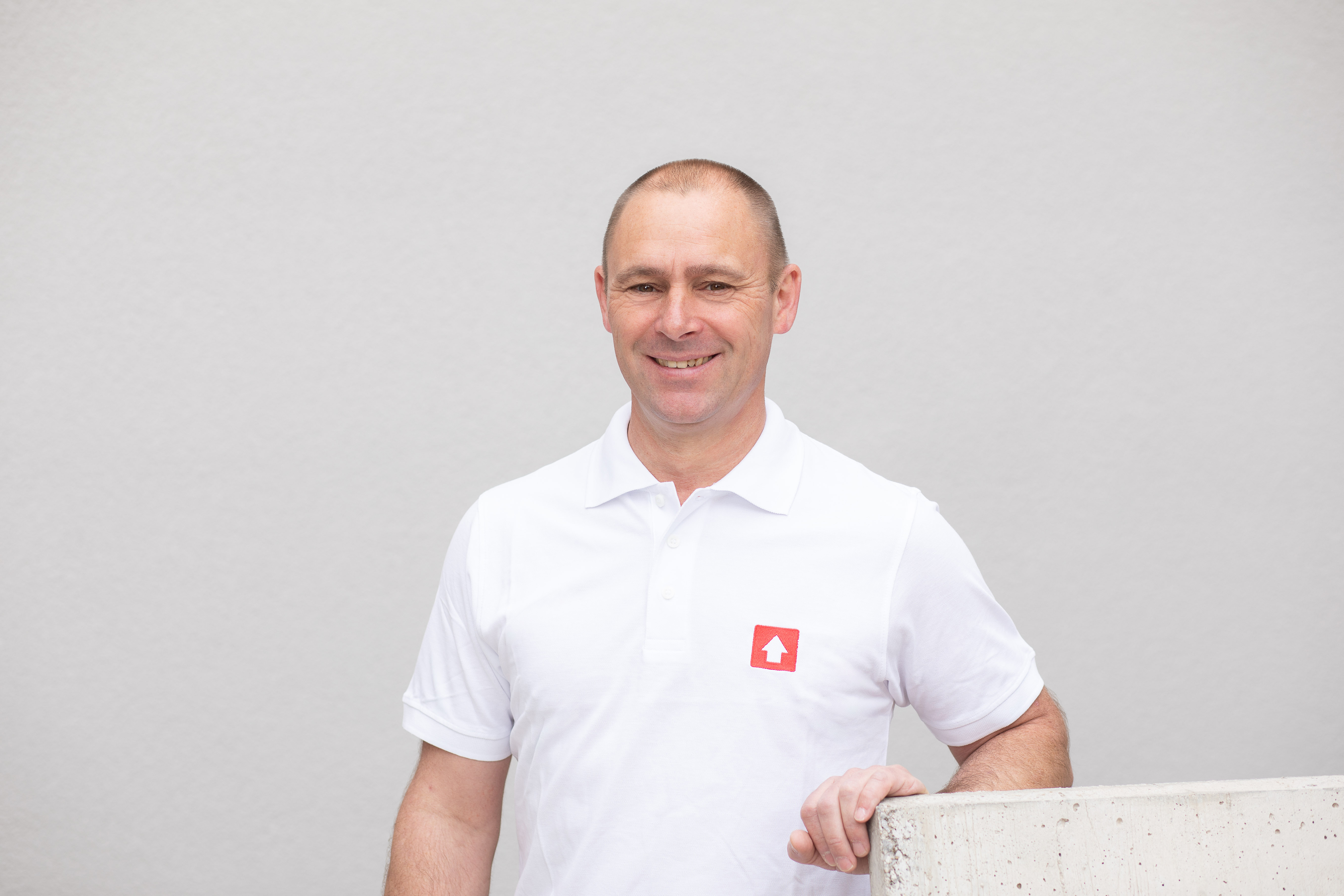 Ein Mann in einem weißen Poloshirt mit einem roten Logo steht vor einer grauen Wand und hält sich an einer Betonfläche fest. Sein Gesicht ist verpixelt.