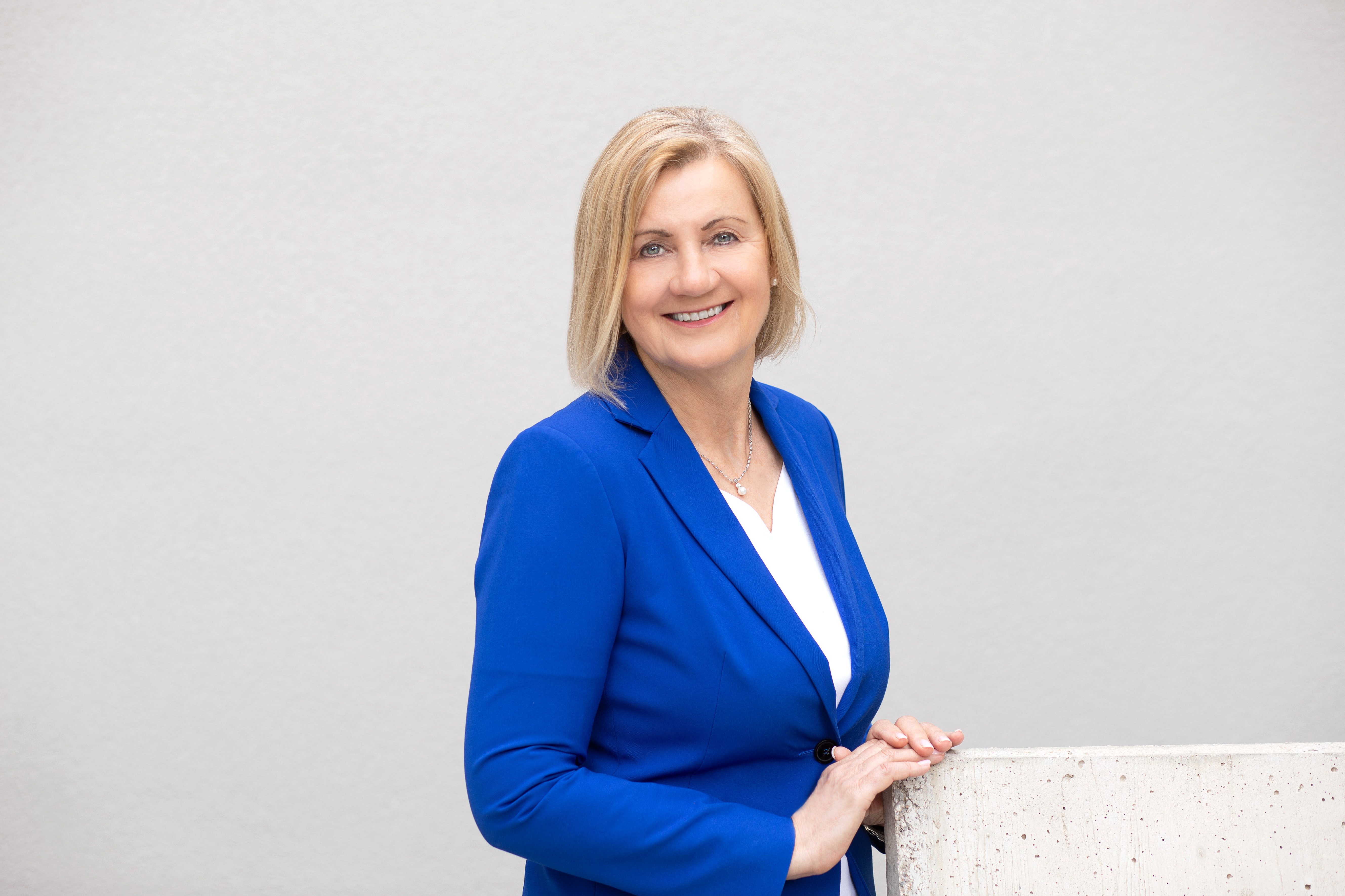 Eine Person mit blondem Haar und verdecktem Gesicht trägt einen blauen Blazer und ein weißes Oberteil. Sie steht vor einem neutralen Hintergrund und stützt sich an einer Betonfläche ab.