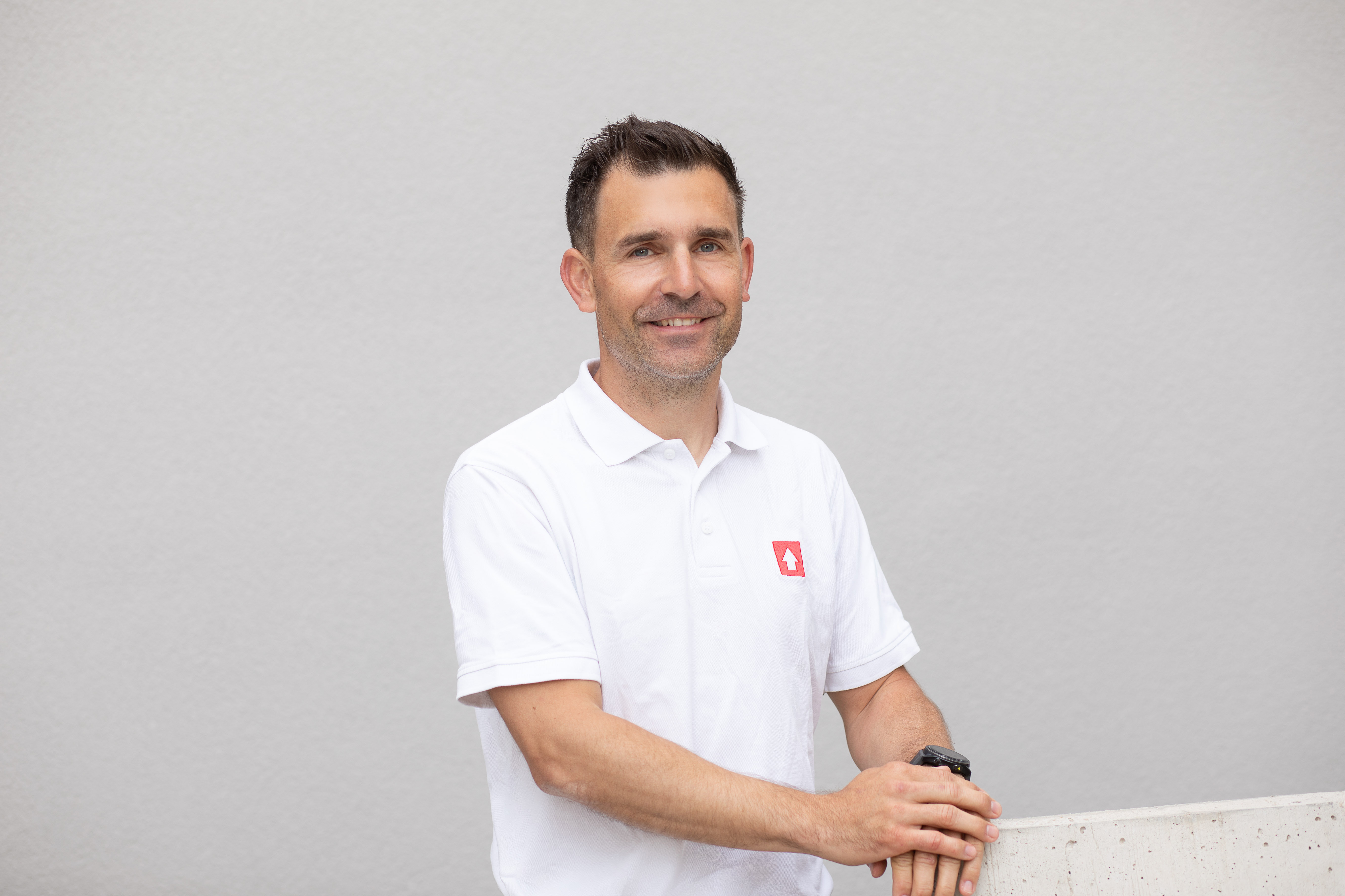 Eine Person in einem weißen Poloshirt mit einem roten Logo stützt sich auf eine Betonbrüstung vor einem neutralen Hintergrund.