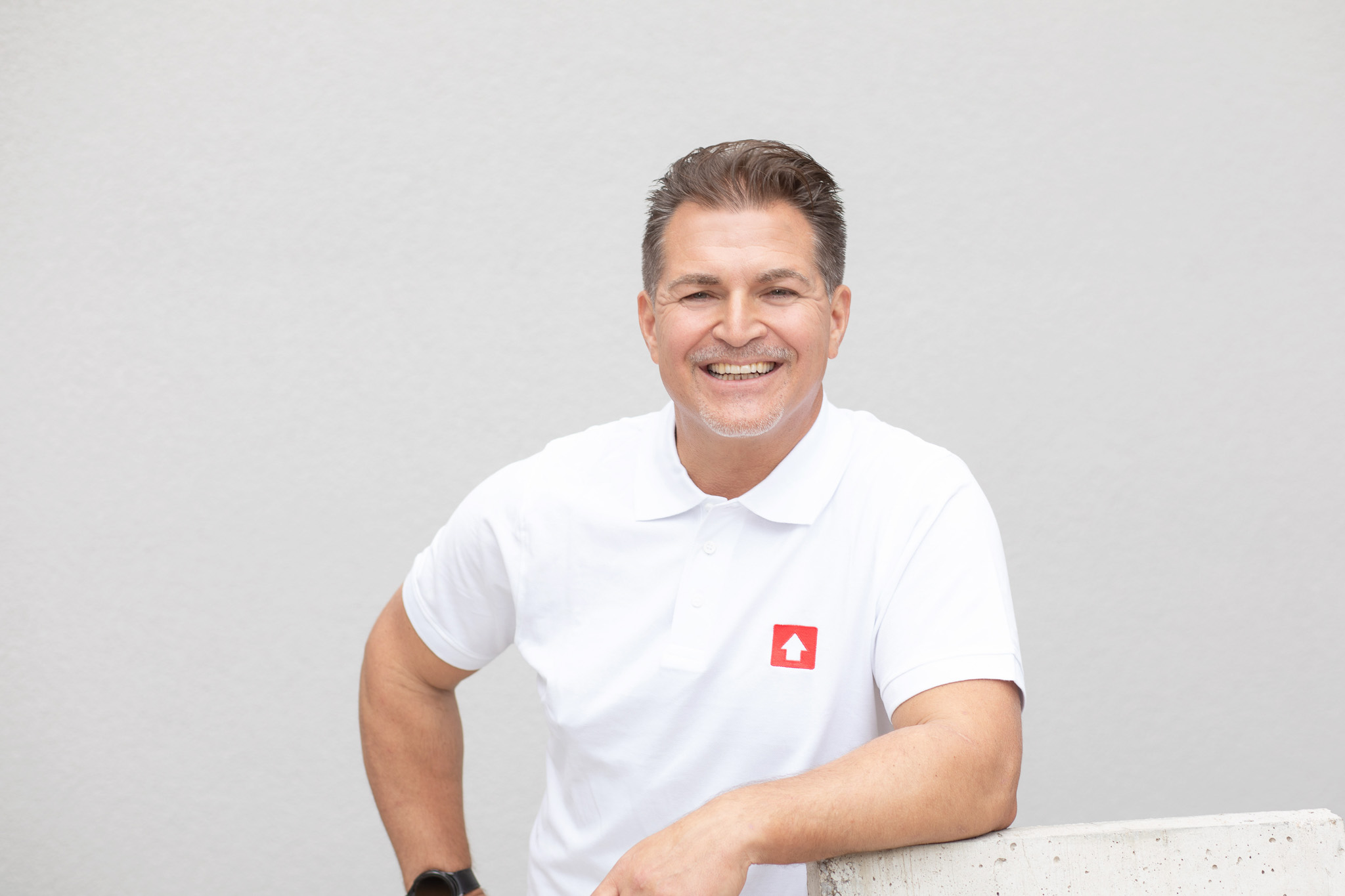 Ein Mann in einem weißen Polo-Shirt mit rotem Logo lehnt an einem Betonblock vor einer grauen Wand. Sein Gesicht ist verpixelt.