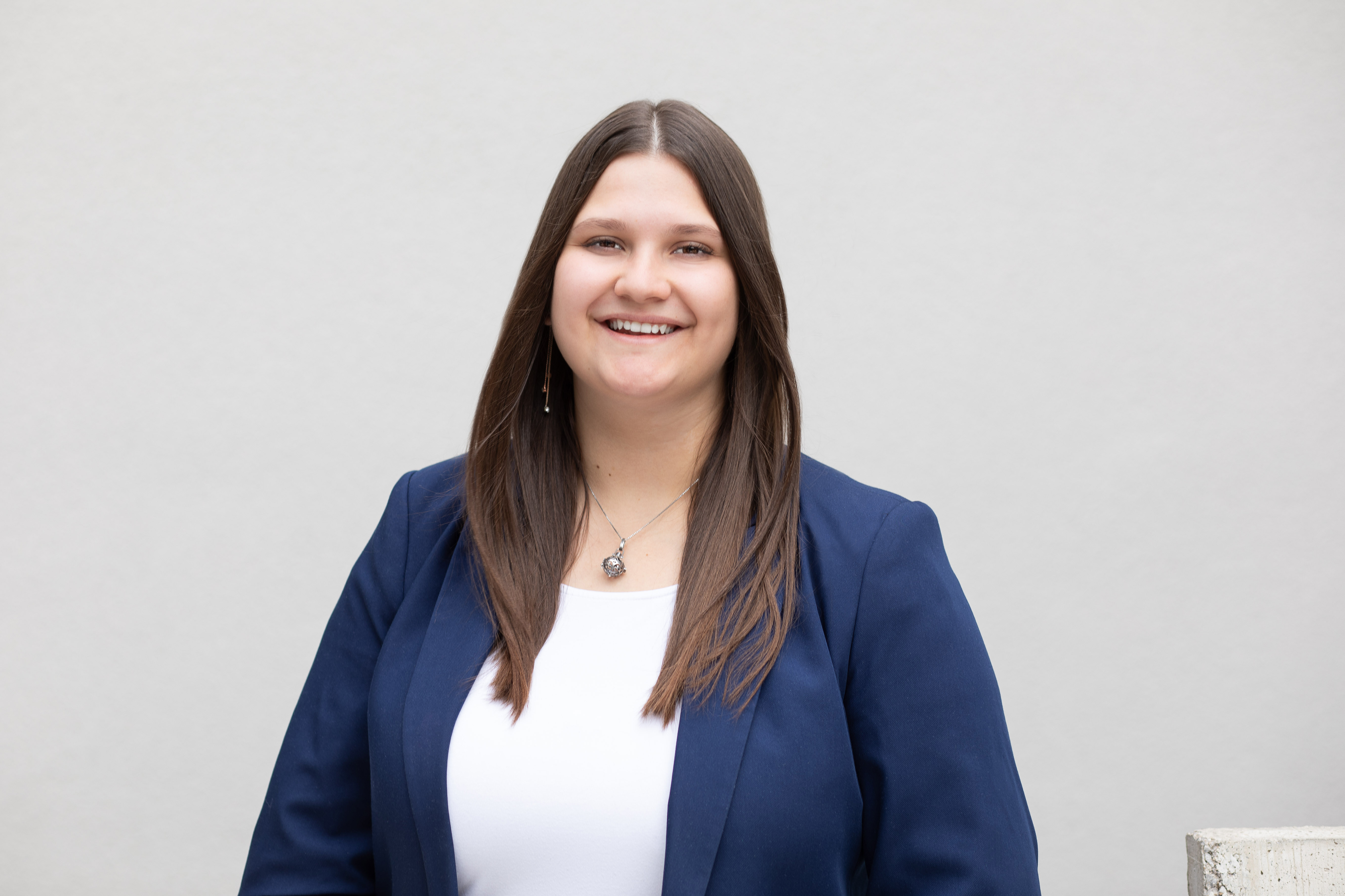 Das Bild zeigt eine Person mit schulterlangem braunem Haar in einem blauen Blazer, weißem Oberteil und einer Halskette, wobei das Gesicht unkenntlich gemacht ist. Der Hintergrund ist grau und schlicht.