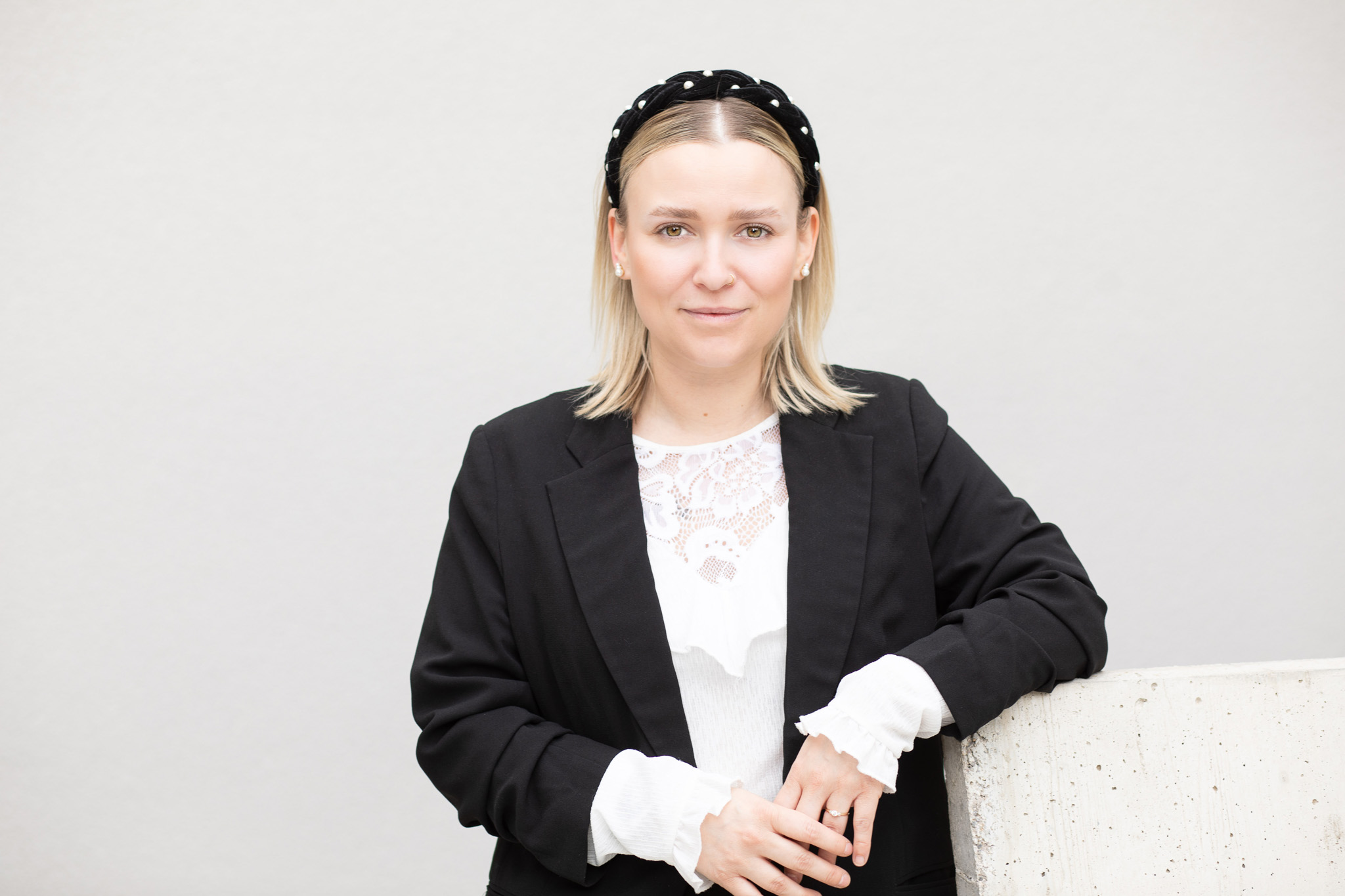 Eine Person in einem schwarzen Blazer und weißem Spitzenoberteil, die sich mit einer entspannten Haltung auf einer Betonkonstruktion abstützt. Der Kopf ist teilweise abgedeckt.