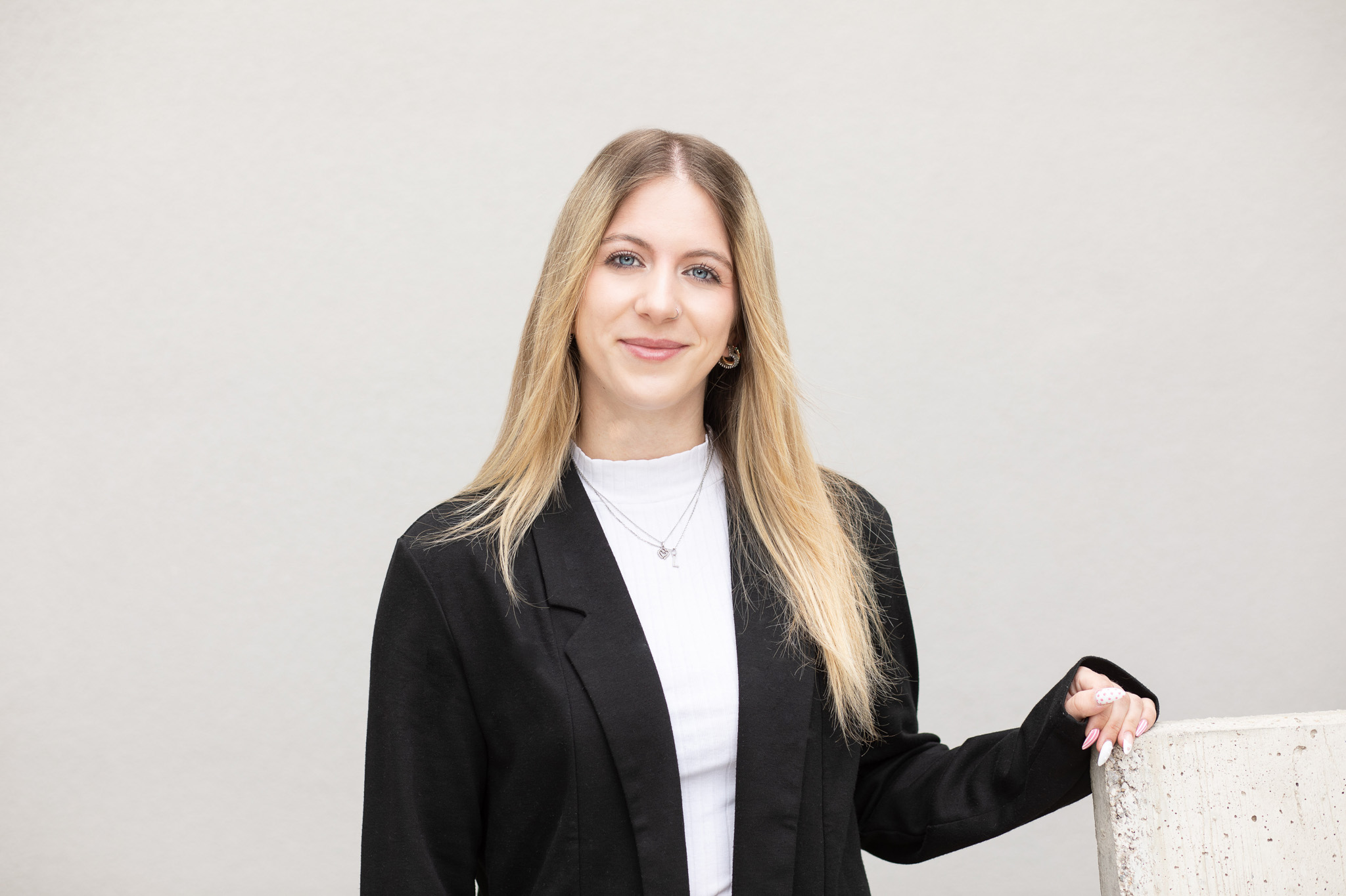 Dieses Bild zeigt eine Person mit langen, blonden Haaren in einem schwarzen Blazer und weißem Oberteil vor einem schlichten Hintergrund. Das Gesicht ist unkenntlich gemacht.