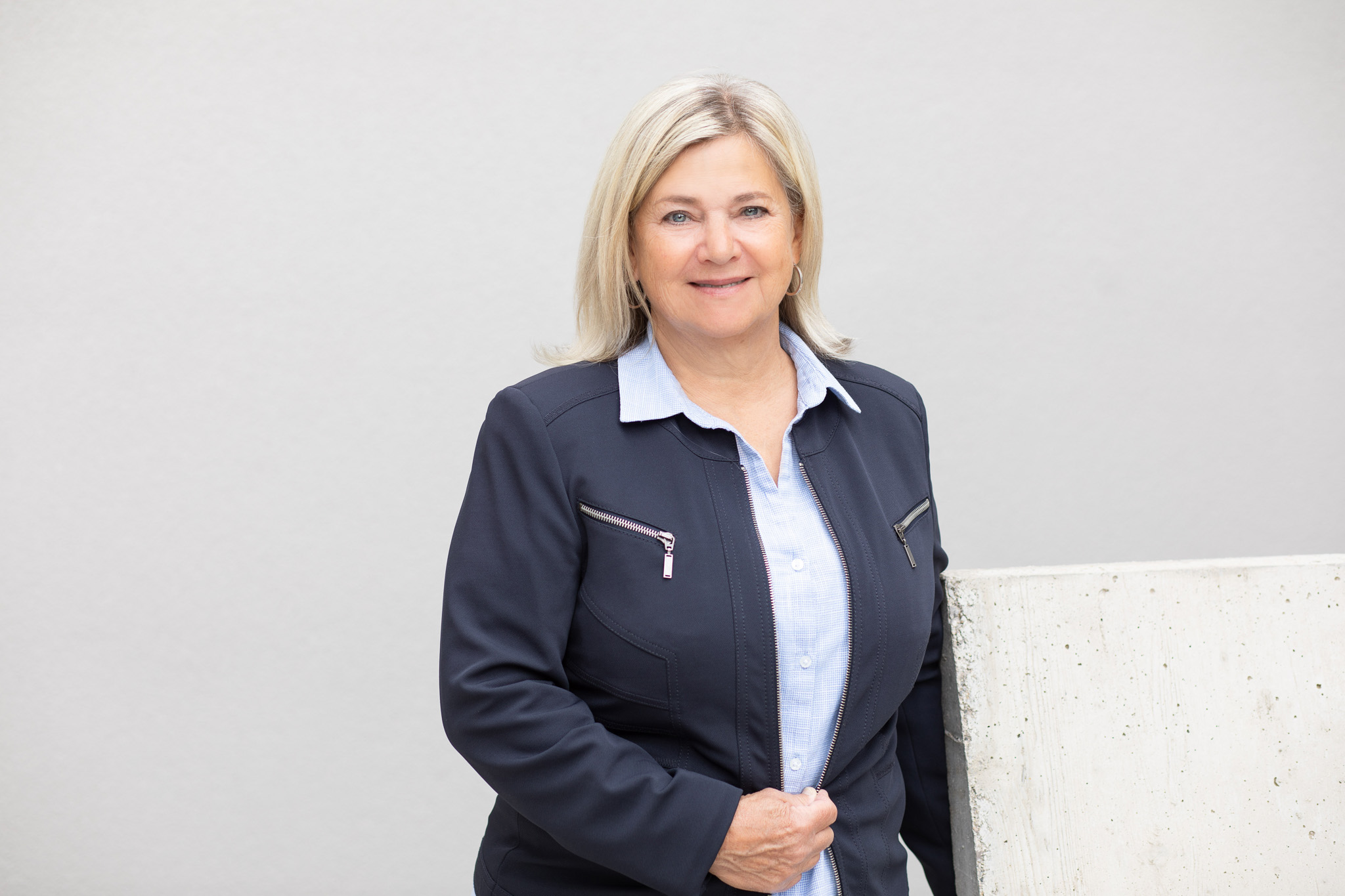 Eine Frau mit blondem Haar steht vor einer grauen Wand. Sie trägt ein dunkelblaues Sakko mit Zippern und eine hellblaue Bluse. Ihre Gesichtszüge sind verdeckt. Die Umgebung wirkt schlicht und neutral.