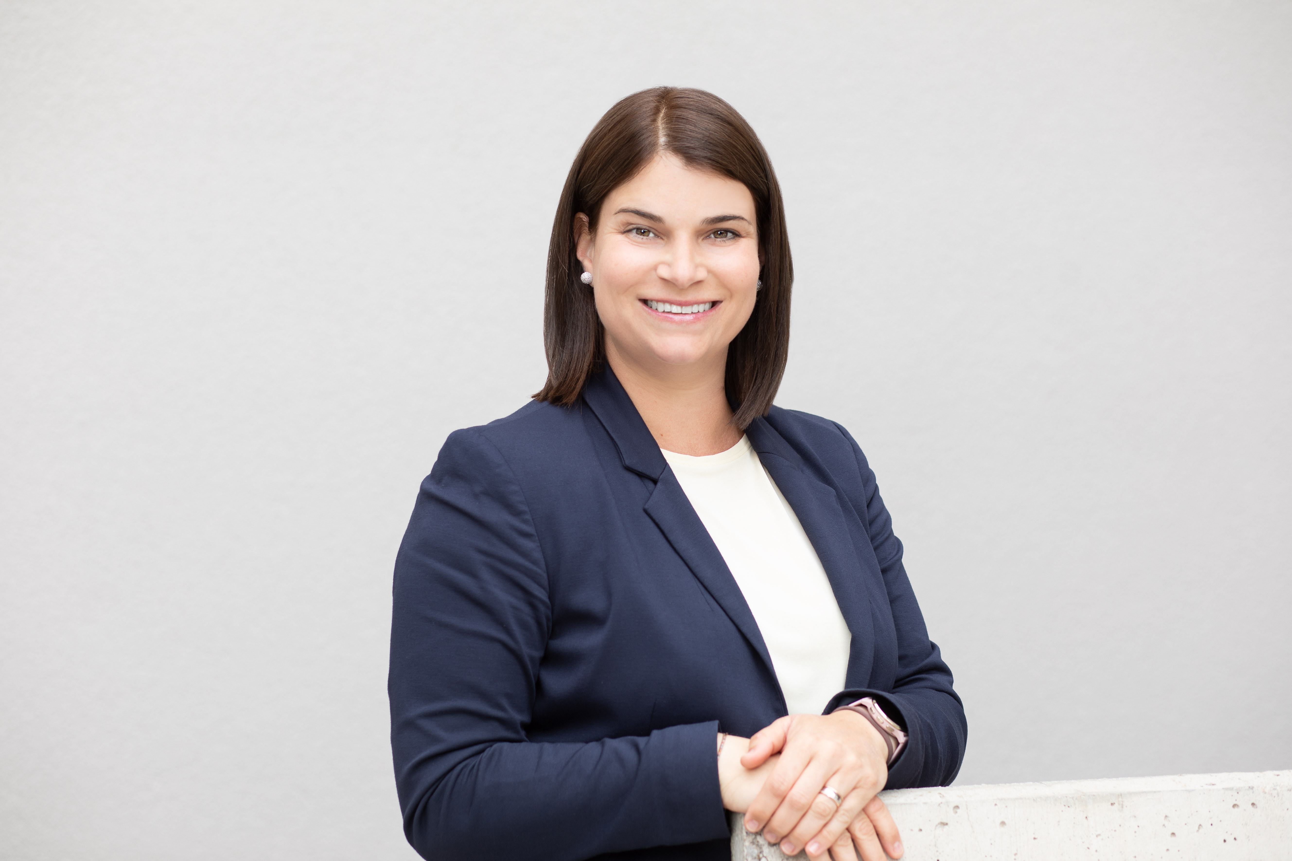 Die Grafik zeigt eine Person mit schulterlangem dunklem Haar in professioneller Kleidung, bestehend aus einem dunkelblauen Blazer und einem weißen Oberteil, die an eine helle Wand lehnt. Ihr Gesicht ist unkenntlich gemacht.