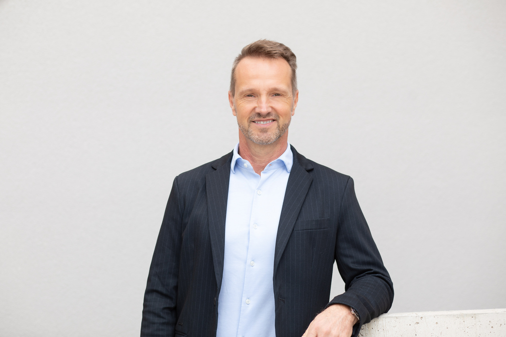 Ein Mann trägt ein Dunkelblaues Sakko mit feiner Nadelstreifenstruktur, ein hellblaues Hemd und eine Uhr am linken Handgelenk. Er steht vor einem neutralen Hintergrund und lehnt an einer hellen Fläche.