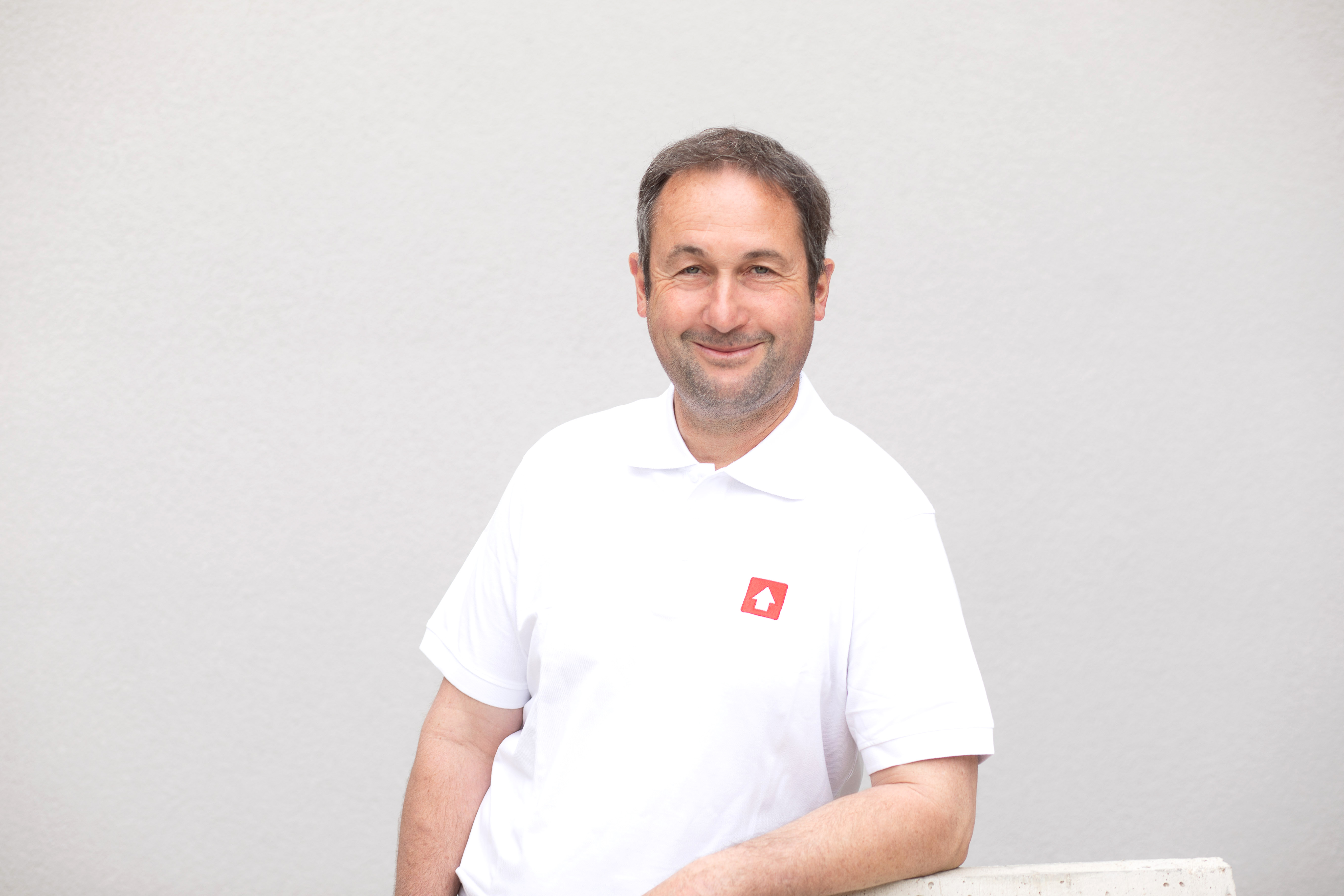 Ein Mann in einem weißen Polohemd mit einem roten Logodruck steht vor einer neutralen, hellgrauen Wand. Sein Gesicht ist unkenntlich gemacht (verpixelt).