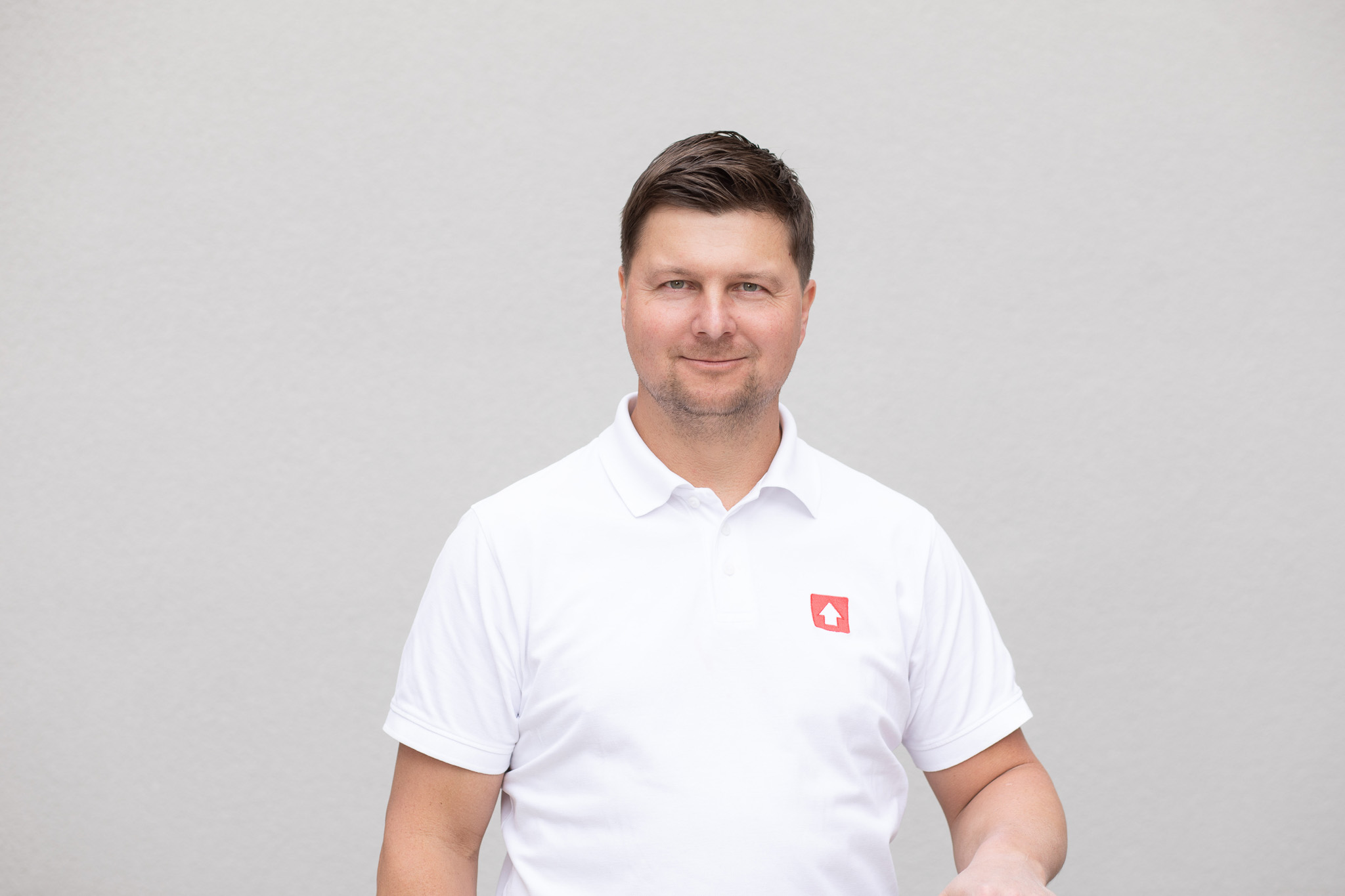 Ein Mann in einem weißen Polohemd mit einem roten Logo auf der linken Brust steht vor einer grauen Hintergrundfläche. Das Gesicht ist unscharf gemacht bzw. zensiert.