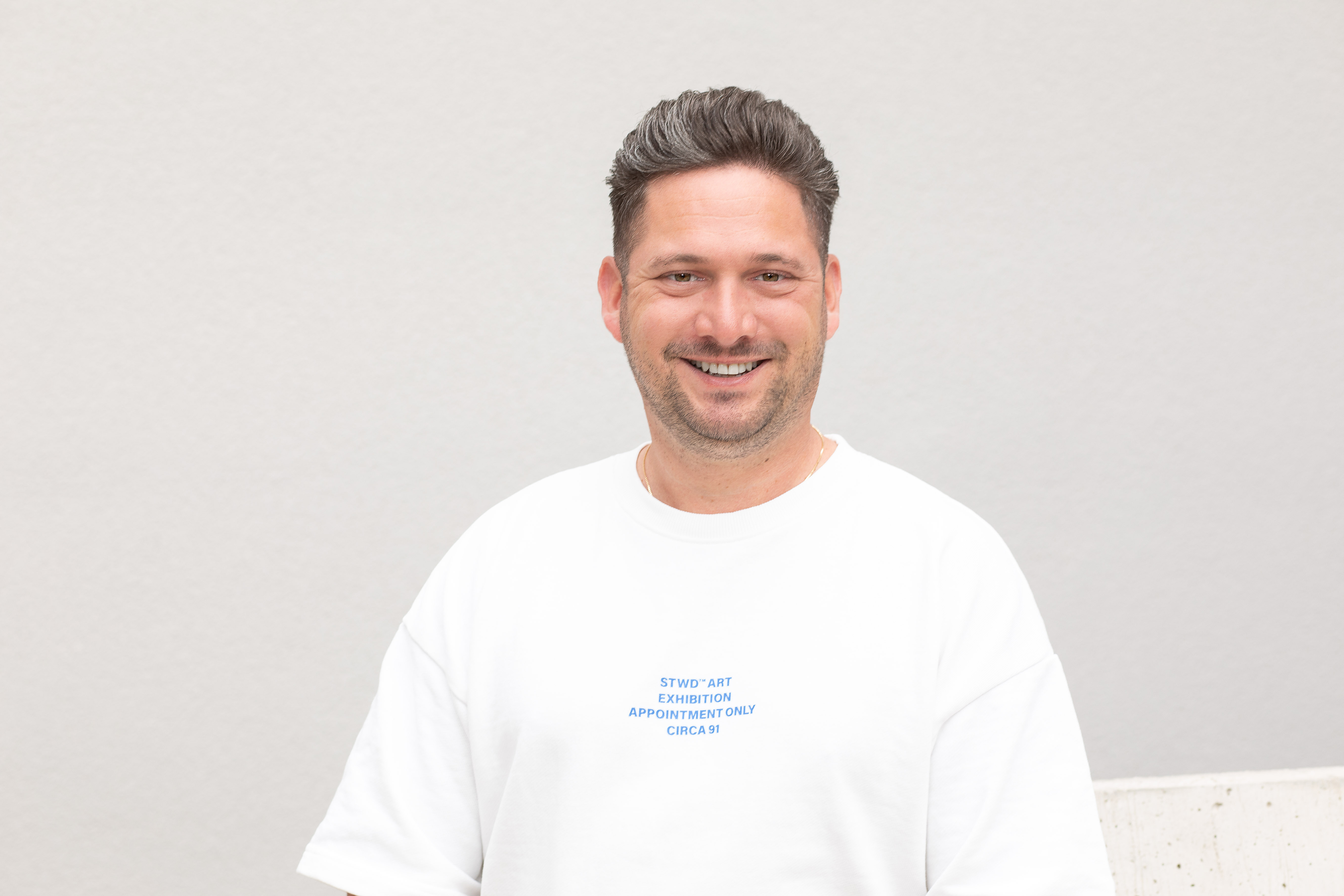 Das Bild zeigt eine Person vor einem hellen Hintergrund, deren Gesicht unkenntlich gemacht wurde. Sie trägt ein weißes Sweatshirt mit einem blauen Text und hat kurze, dunkle Haare.