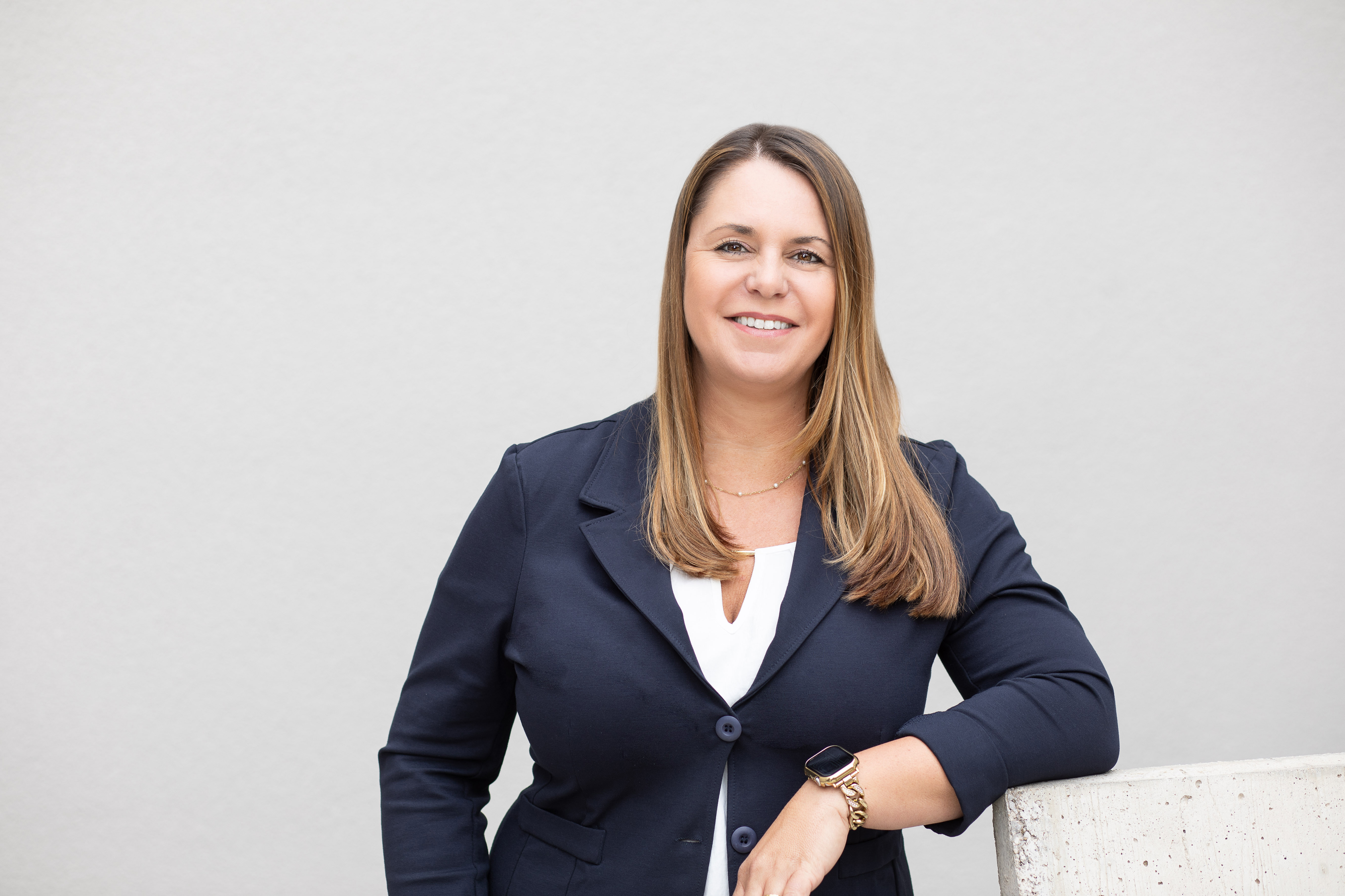 Diese Grafik zeigt eine Person mit blondem, glattem Haar in einem formellen dunkelblauen Blazer auf neutralem Hintergrund. Das Gesicht wurde unkenntlich gemacht. Sie trägt Schmuck und eine Smartwatch.